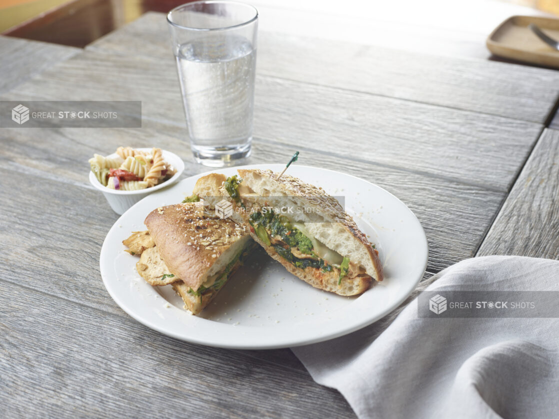 Grilled chicken panini sandwich with spinach, pesto, cheese and sauce on white plate topped with parmesan cheese with a side pasta salad and glass of water on a wood table