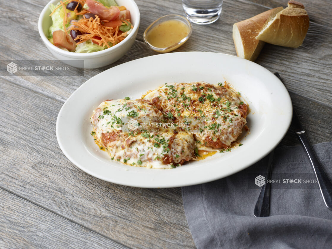 Two chicken parmesan cutlets on a white plate with a side salad and fresh bread in the background on a wooden table