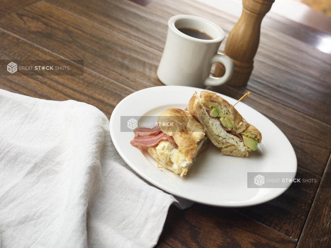 Breakfast sandwich with cheese, avocado, bacon and egg whites on a croissant with a cup of black coffee in the background on a wooden table