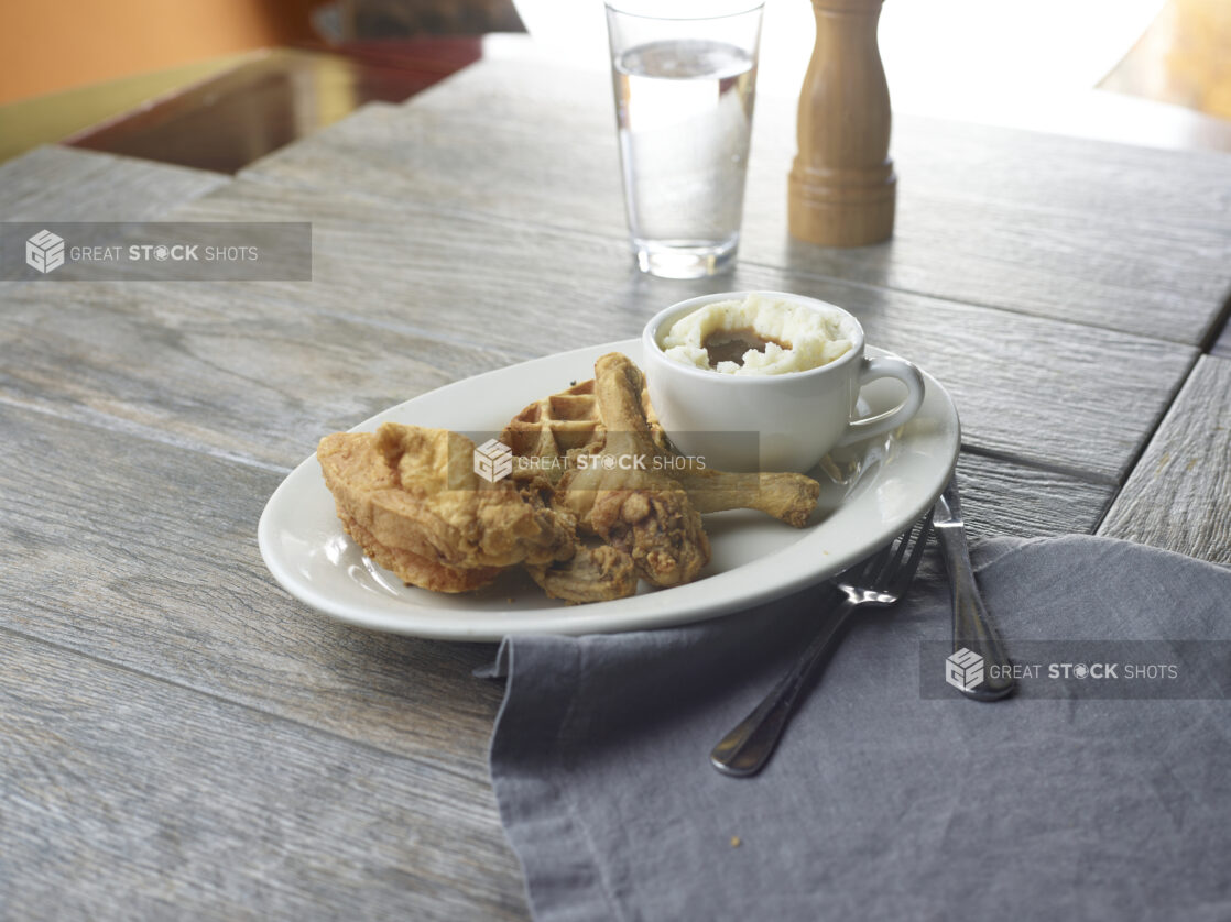 Fried chicken and a waffle on a pate with a side of mashed potatoes and gracy in a mug on a wooden table