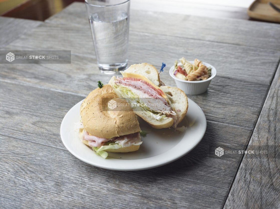 Turkey, ham and roast beef sandwich with swiss and cheddar cheese, lettuce, tomato and mayonnaise on a poppyseed bun with a side pasta salad on a wood table