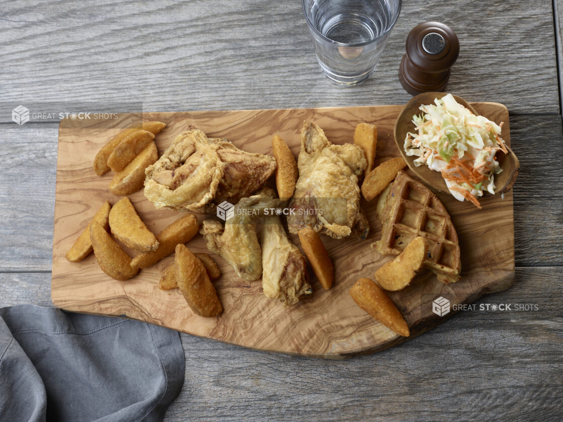 Overhead of fried chicken, potato wedges and waffle on a live edge wooden board with a side of coleslaw on a wooden table