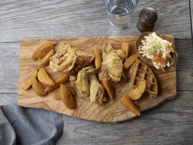 Overhead of fried chicken, potato wedges and waffle on a live edge wooden board with a side of coleslaw on a wooden table