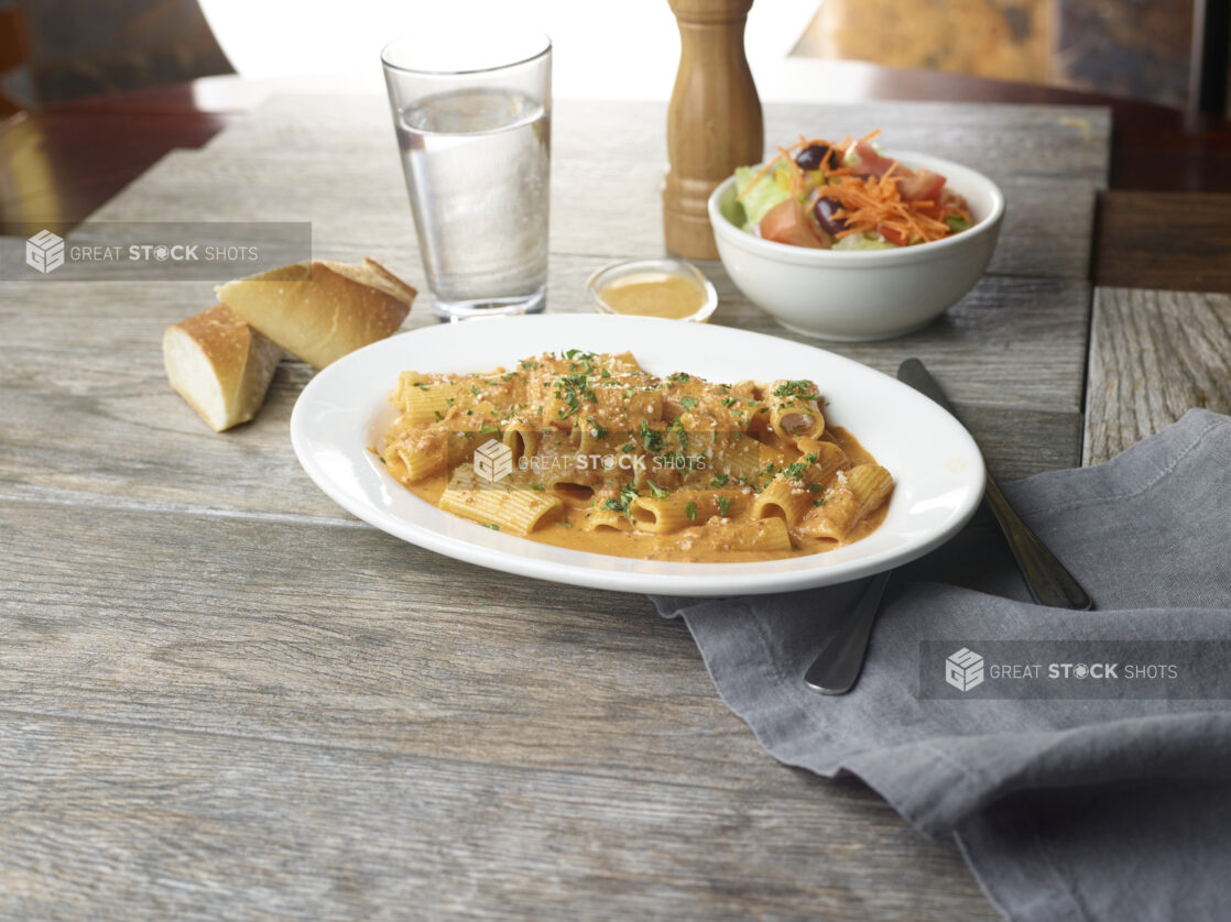 Plate of rigatoni in a rosé sauce topped with grated parmesan and fresh parsley with a side salad and fresh bread in the background on a wooden table