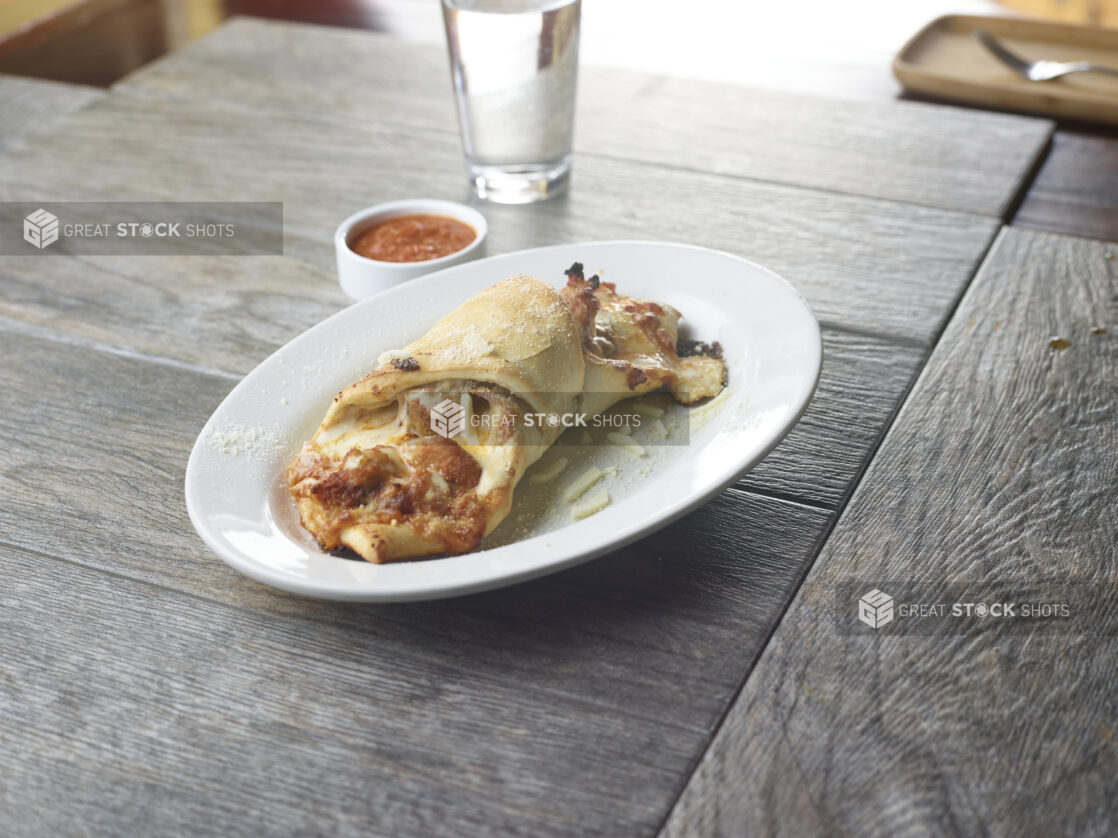 A cheese and tomato sauce calzone on a white plate with a ramekin of marinara sauce and a glass of water in the background