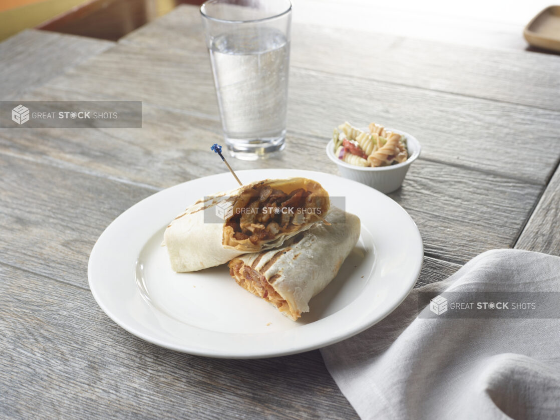 A sandwich wrap / burrito, halved, on a white plate beside a ramekin of pasta salad and a glass of water