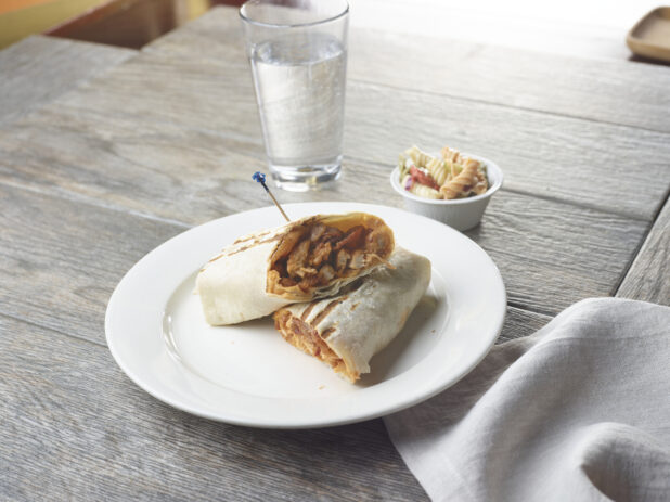 A sandwich wrap / burrito, halved, on a white plate beside a ramekin of pasta salad and a glass of water