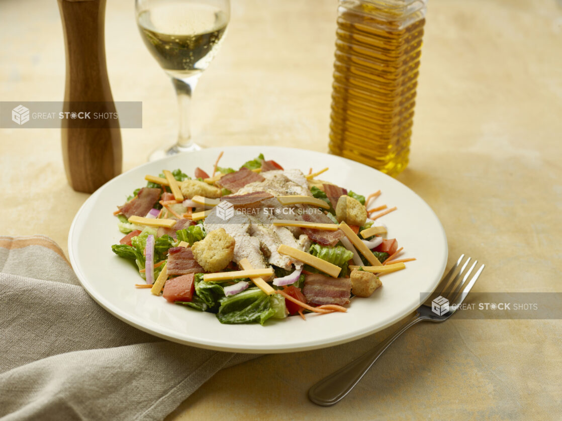 Salad with bacon, chicken, red onion, croutons, red pepper and shredded cheddar with a glass of white wine on a beige background