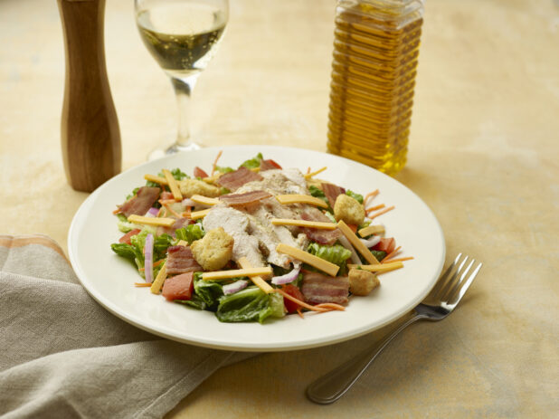 Salad with bacon, chicken, red onion, croutons, red pepper and shredded cheddar with a glass of white wine on a beige background