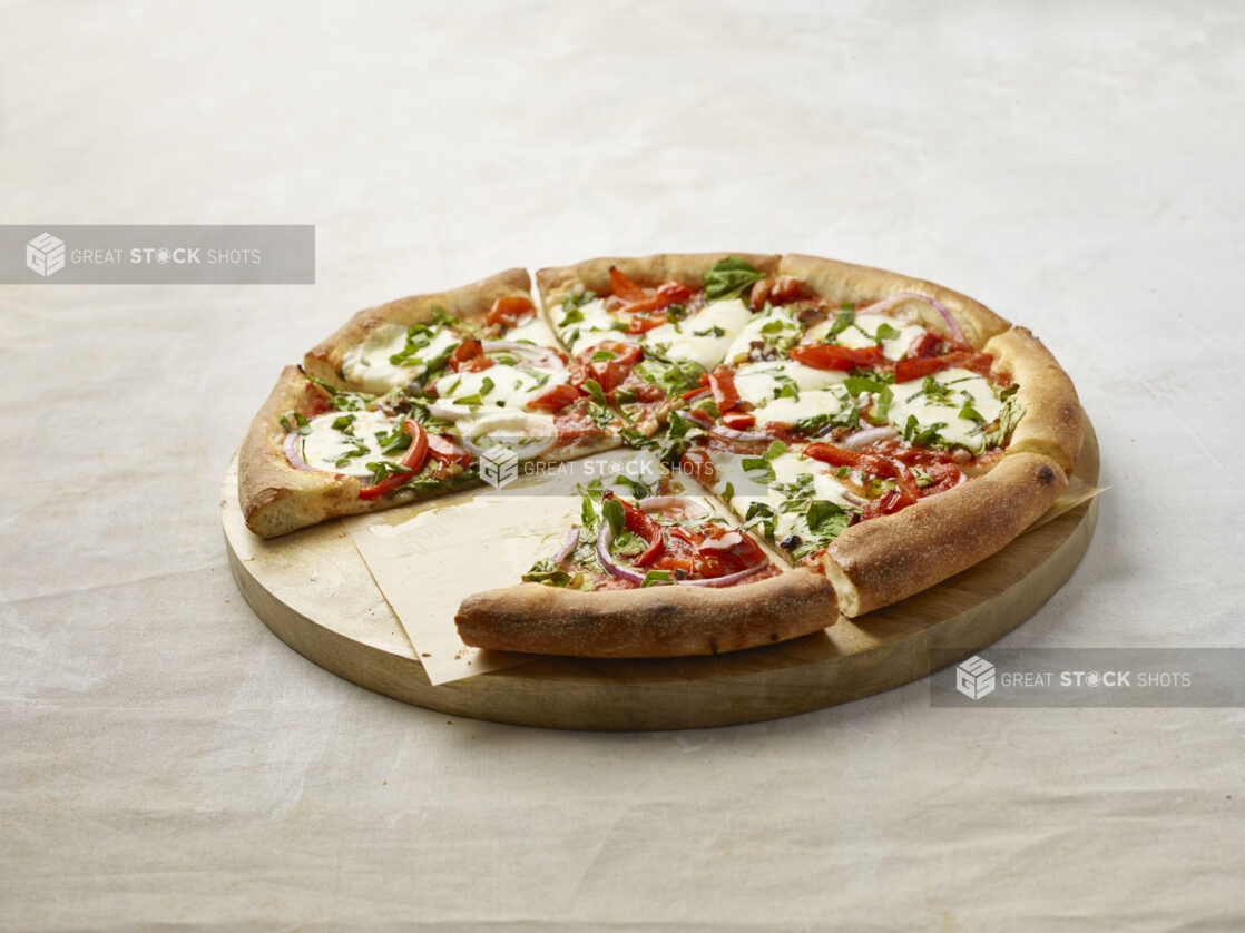 Sliced pizza with red pepper, spinach and onion topped with fresh basil with a slice missing on a wooden board with parchment paper