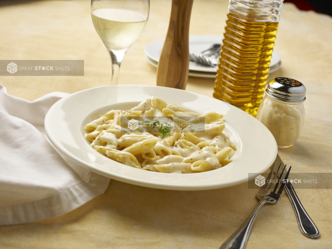 Bowl of Alfredo penne pasta with a glass of white wine on a yellow background