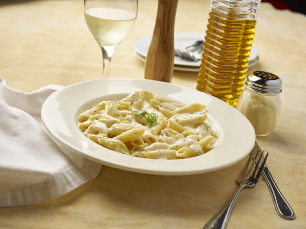 Bowl of Alfredo penne pasta with a glass of white wine on a yellow background