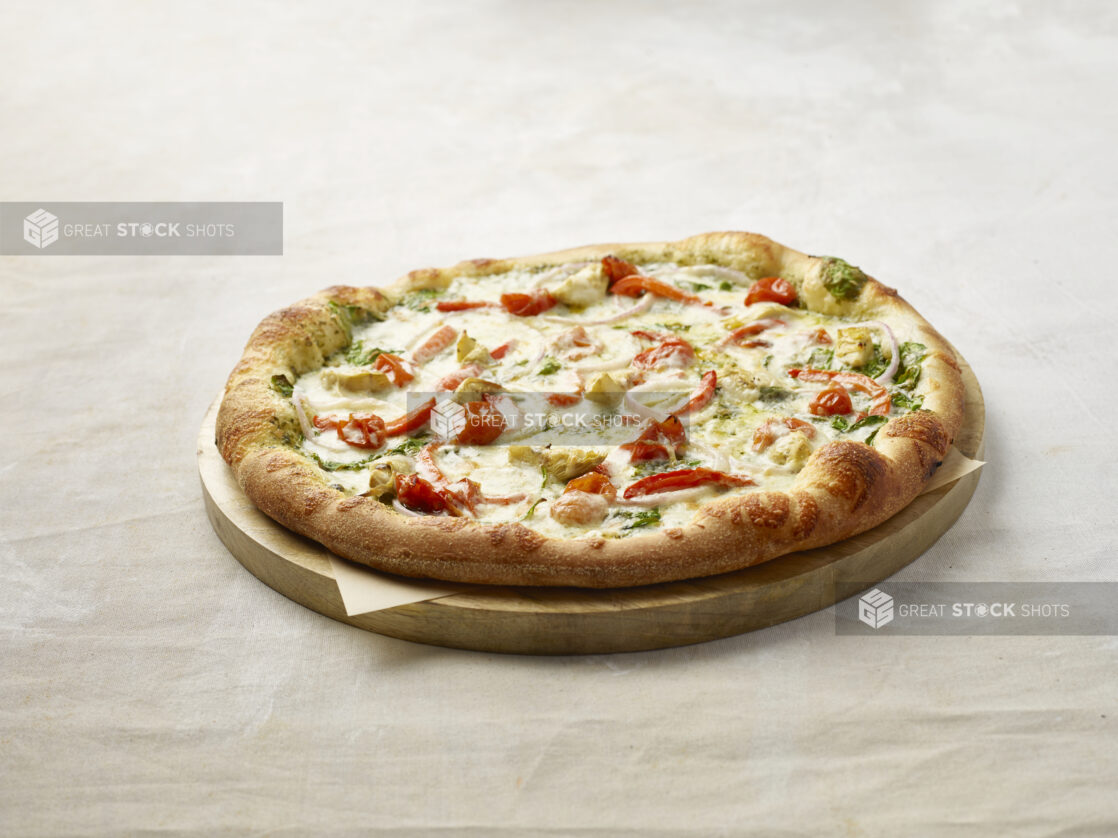 Whole pizza with cherry tomatoes, red pepper, artichoke, spinach and onion with a pesto base on a wooden board with parchment paper