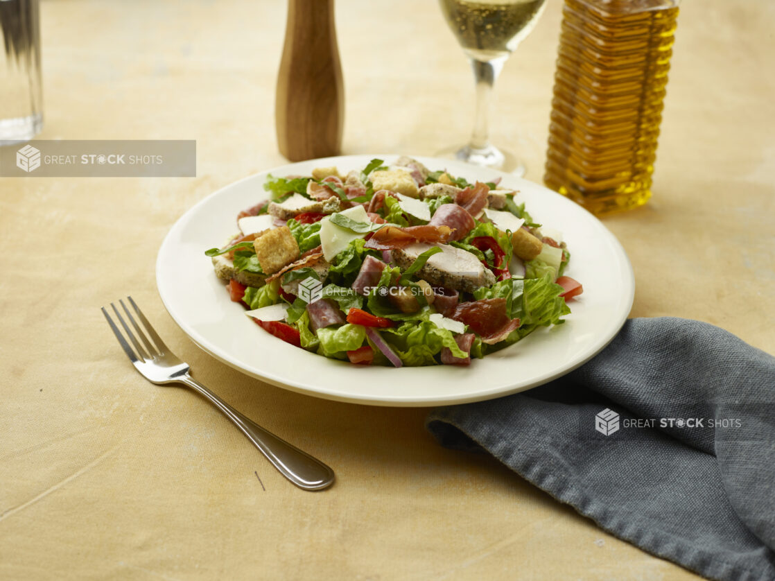 Salad with prosciutto, chicken, salami, red onion, red peppers and shaved parmesan with a glass of white wine on a beige background