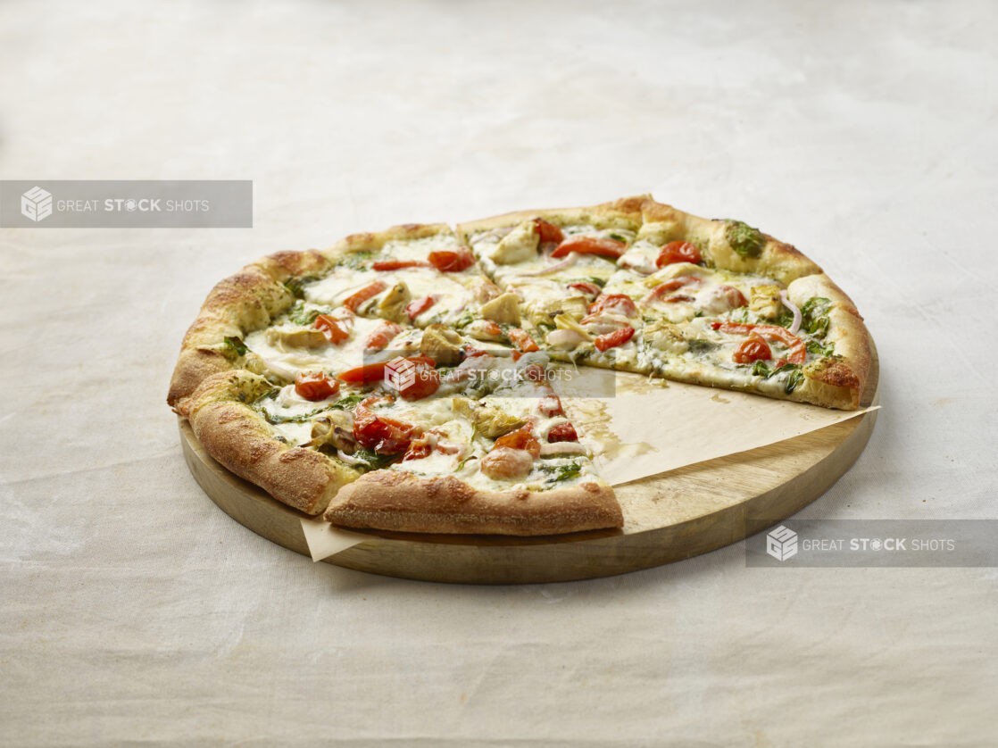 Sliced pizza with cherry tomatoes, red pepper, artichoke, spinach and onion with a pesto base with a slice missing on a wooden board with parchment paper