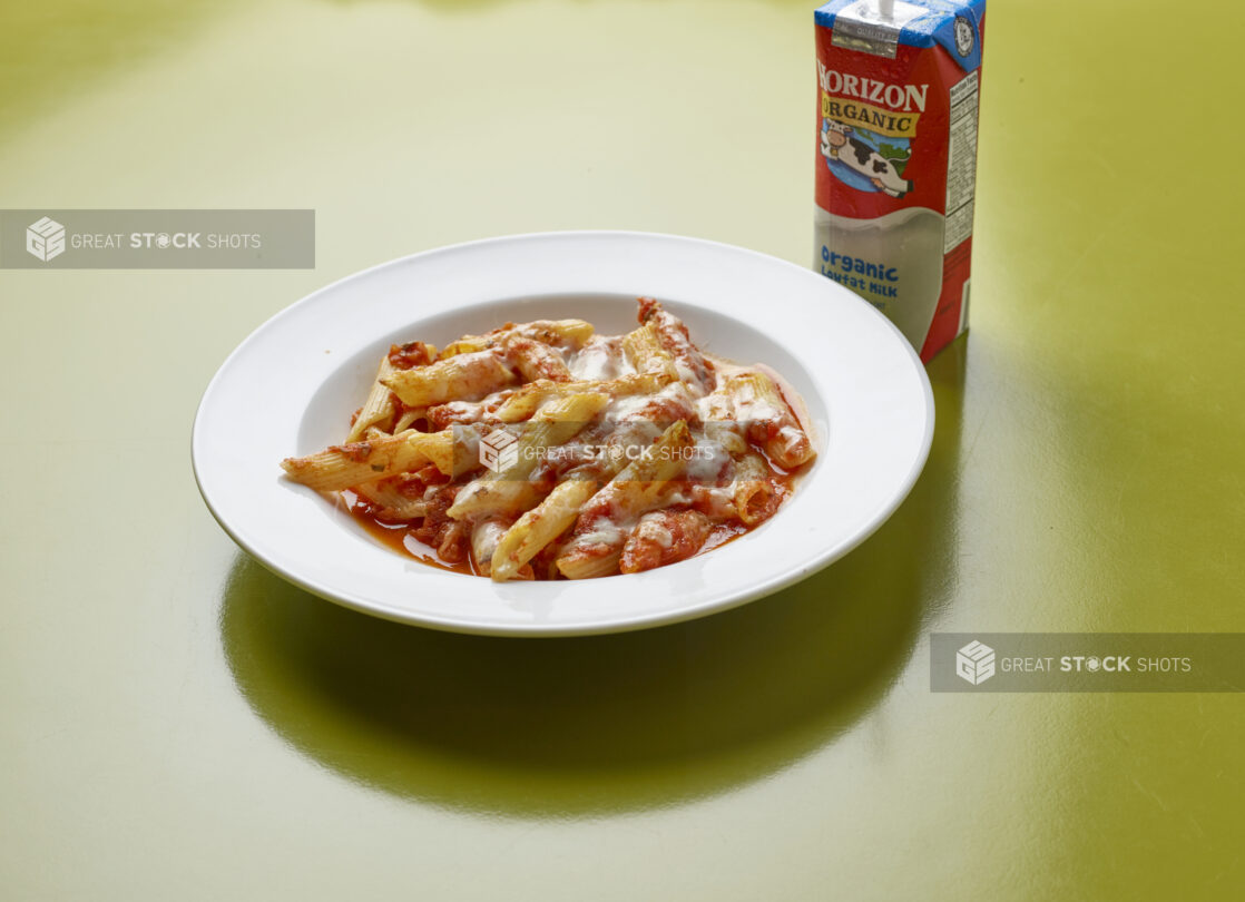 Kids meal of penne with tomato sauce, topped with melted cheese with drink box of milk on a green background