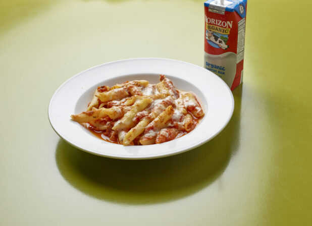 Kids meal of penne with tomato sauce, topped with melted cheese with drink box of milk on a green background