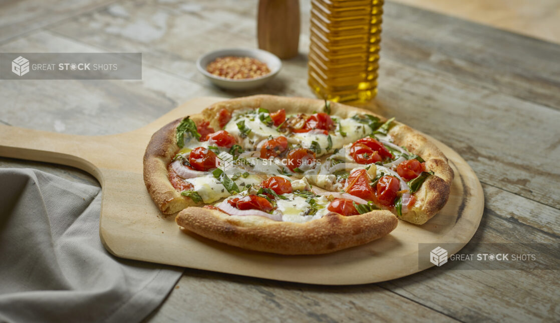 Sliced pizza with cherry tomatoes, onion and fresh basil on a paddle with toppings in the background