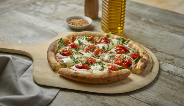 Sliced pizza with cherry tomatoes, onion and fresh basil on a paddle with toppings in the background