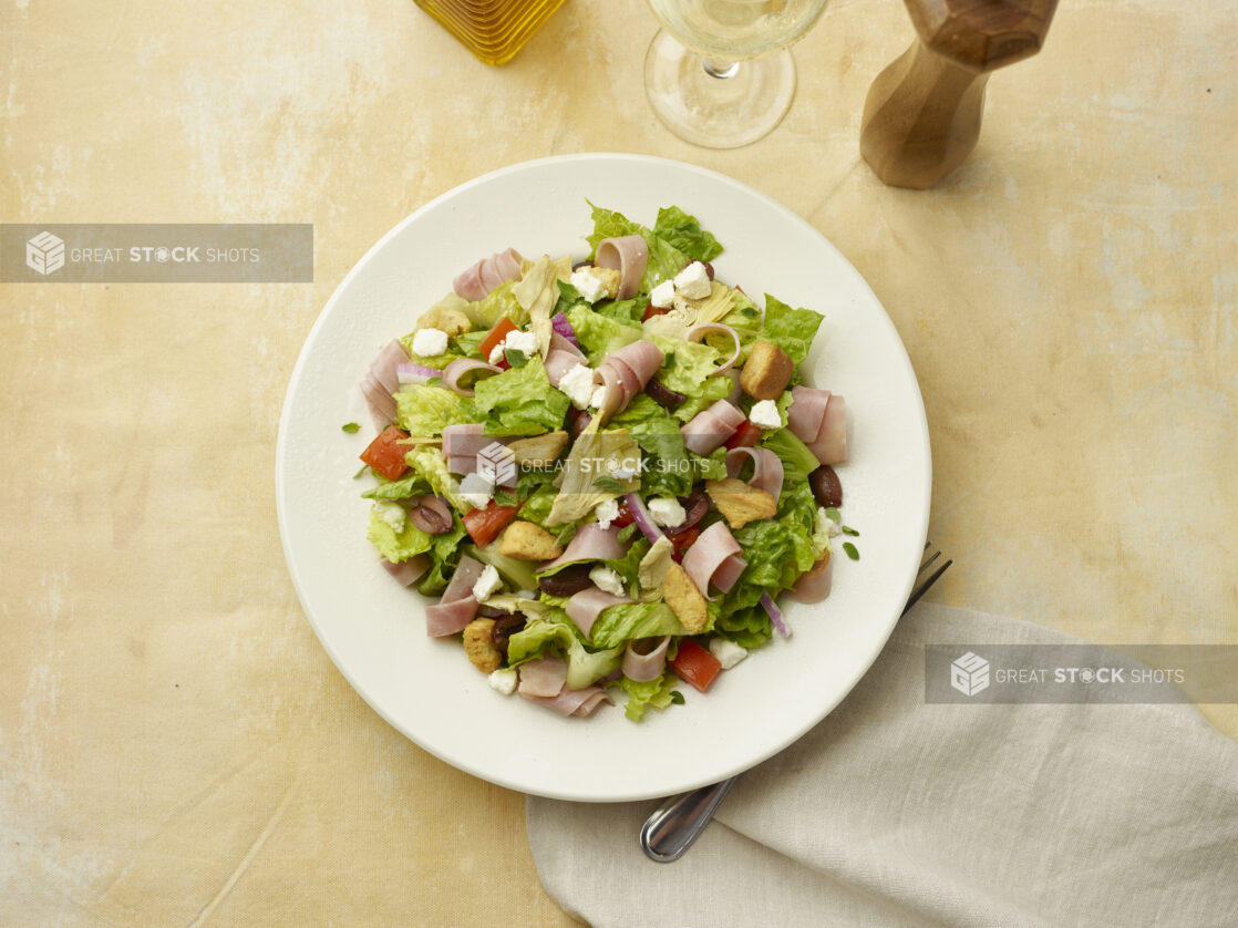 Overhead view of salad with sliced ham, feta, red pepper, croutons, red onion and black olives with a glass of white wine on a beige background