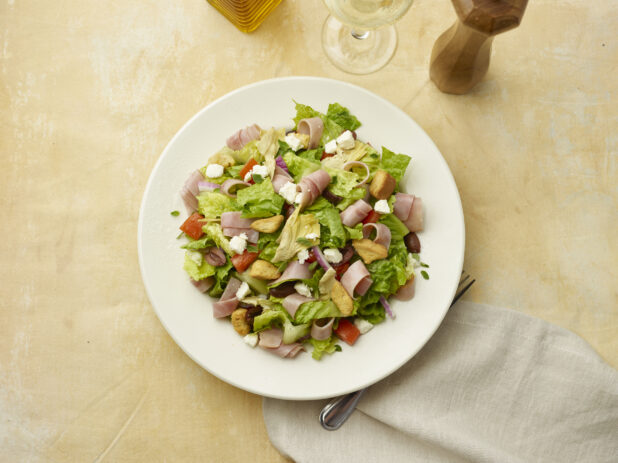 Overhead view of salad with sliced ham, feta, red pepper, croutons, red onion and black olives with a glass of white wine on a beige background