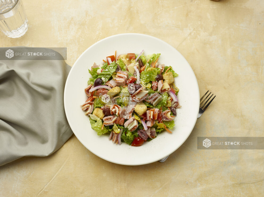 Overhead view of salad with sliced ham, salami and provolone squares on top of red onion, black olives and croutons on a beige background