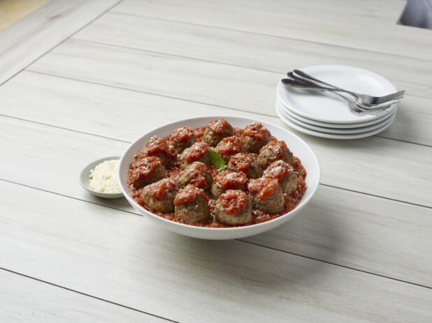 Large bowl of meatballs in tomato sauce with a stack of white dishes and cutlery in the background