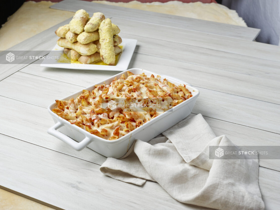 Baked tomato penne pasta with an order of garlic bread sticks on a light wooden background