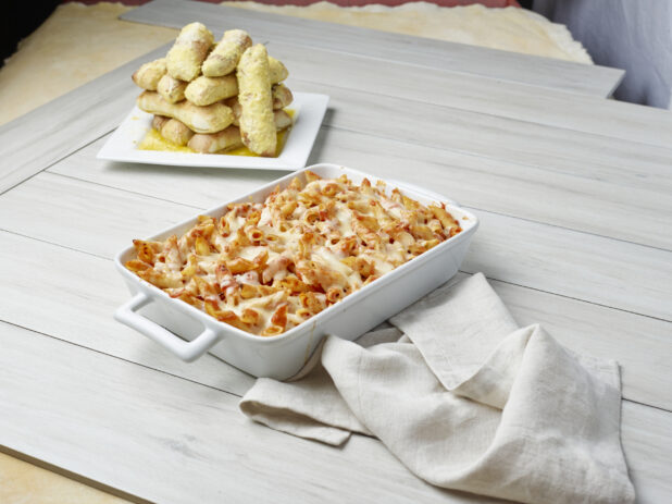 Baked tomato penne pasta with an order of garlic bread sticks on a light wooden background