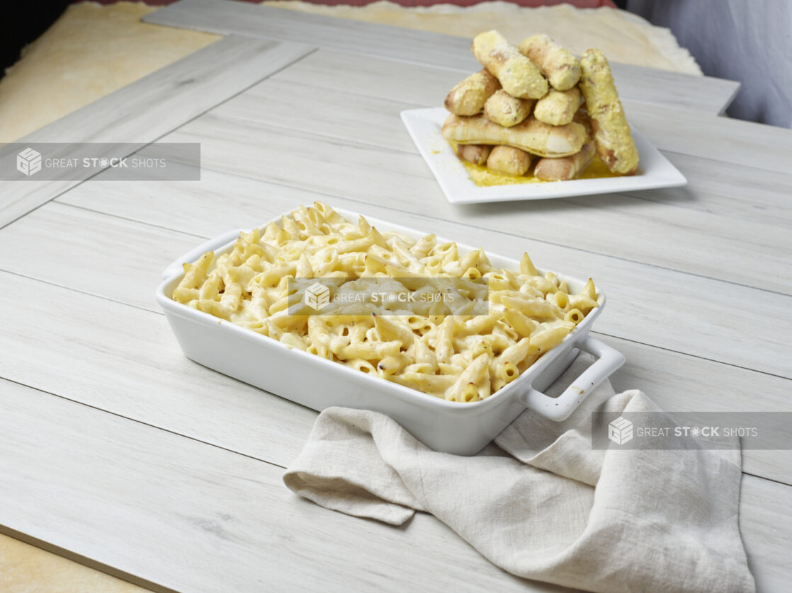 Baked cheesy Alfredo penne pasta with an order of garlic bread sticks on a light wooden backgroundh