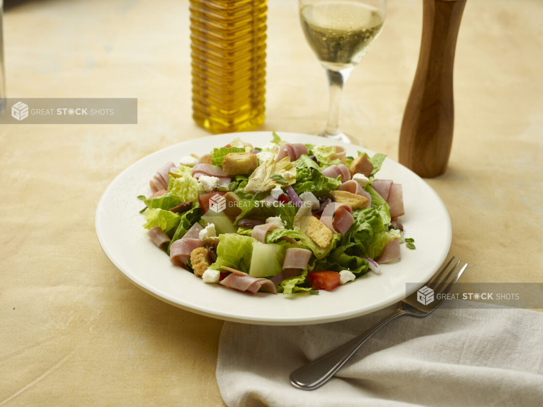 Salad with sliced ham, feta, red pepper, croutons, red onion and black olives with a glass of white wine on a beige background