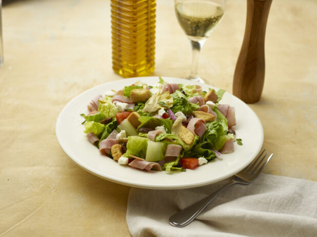 Salad with sliced ham, feta, red pepper, croutons, red onion and black olives with a glass of white wine on a beige background
