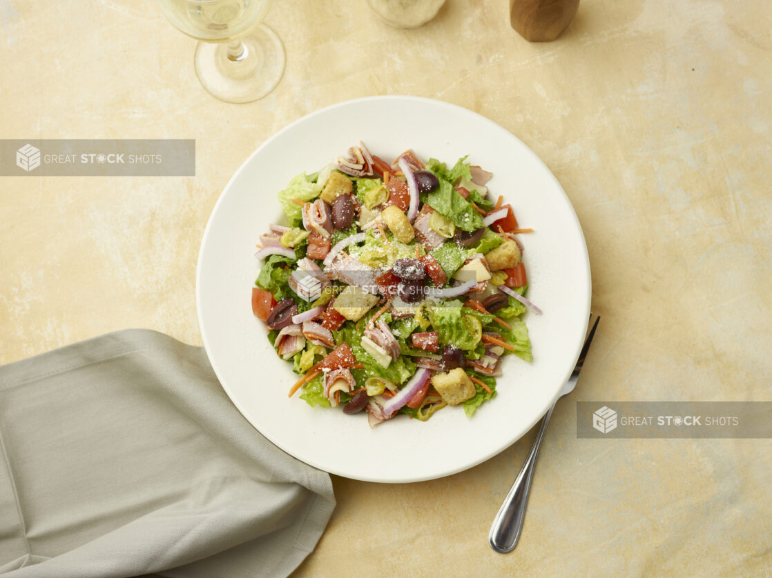 Overhead view of salad with sliced ham, salami and provolone squares on top of red onion, black olives and croutons with a glass of white wine on a beige background