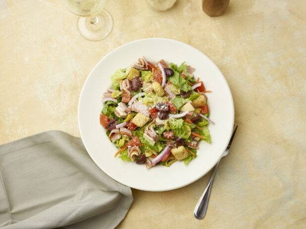 Overhead view of salad with sliced ham, salami and provolone squares on top of red onion, black olives and croutons with a glass of white wine on a beige background