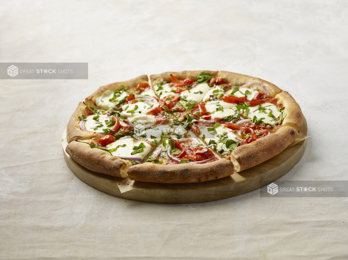 Whole Sliced New York Style Pizza with Roasted Red Peppers, Red Onions, Fresh Basil and Fresh Mozzarella Cheese on a Wooden Platter on a Light Beige Surface and Background