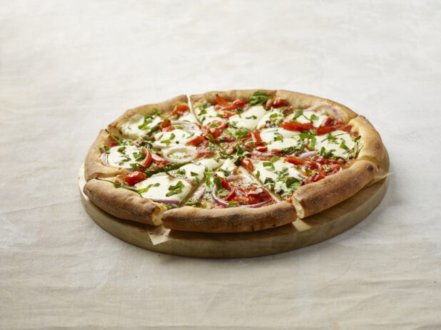 Whole Sliced New York Style Pizza with Roasted Red Peppers, Red Onions, Fresh Basil and Fresh Mozzarella Cheese on a Wooden Platter on a Light Beige Surface and Background