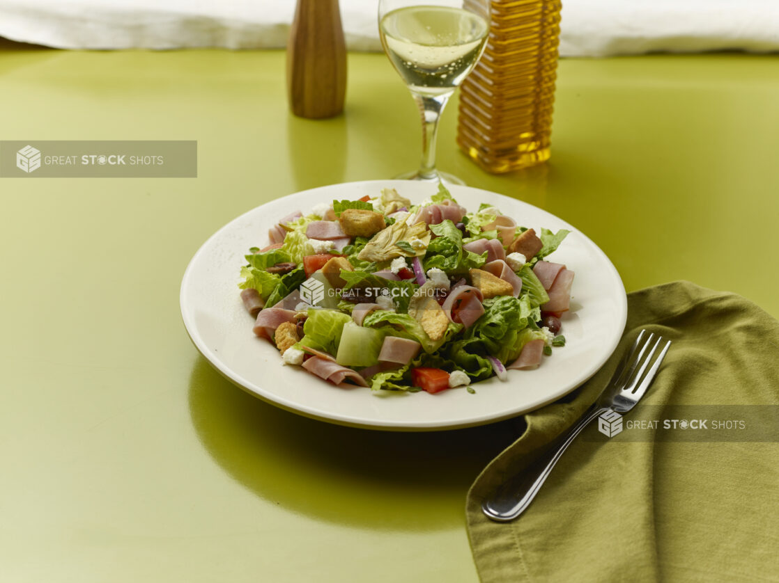 Chopped salad with sliced ham, feta, red pepper, croutons, red onion and black olives with a glass of white wine on a green background