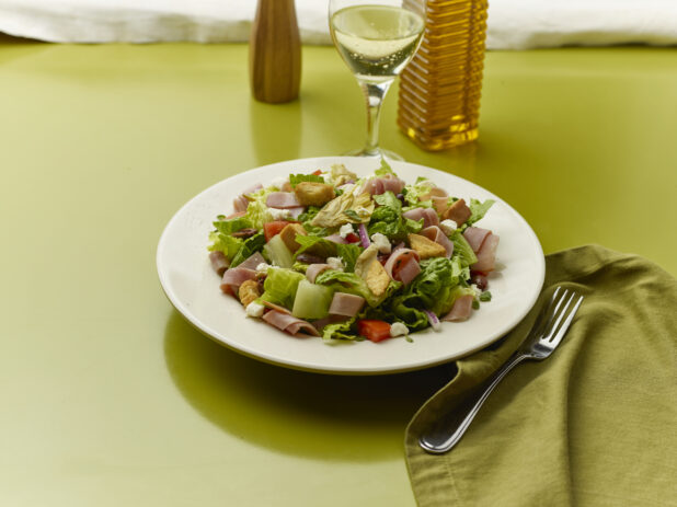 Chopped salad with sliced ham, feta, red pepper, croutons, red onion and black olives with a glass of white wine on a green background
