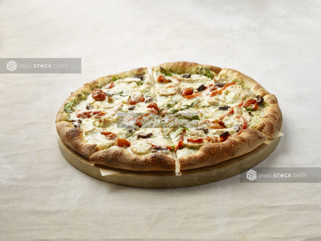 Sliced pizza with chicken, cherry tomatoes, black olives and red pepper with a pesto base on top on a wooden board with parchment paper