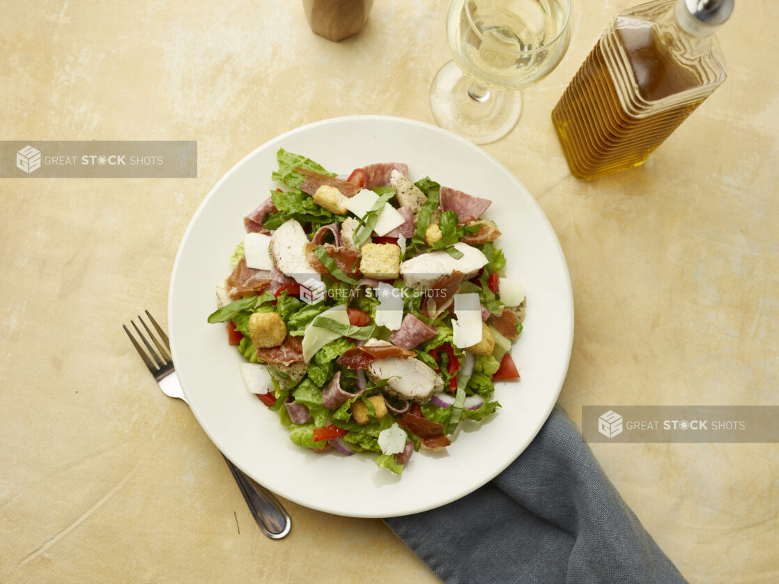 Overhead view of salad with prosciutto, chicken, salami, red onion, red peppers and shaved parmesan with a glass of white wine on a beige background