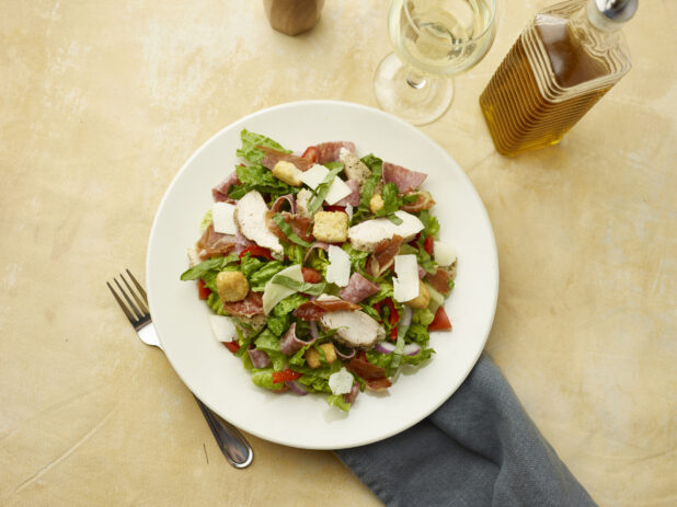 Overhead view of salad with prosciutto, chicken, salami, red onion, red peppers and shaved parmesan with a glass of white wine on a beige background