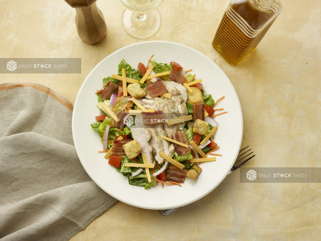 Overhead view of salad with bacon, chicken, red onion, croutons, red pepper and shredded cheddar with a glass of white wine on a beige background