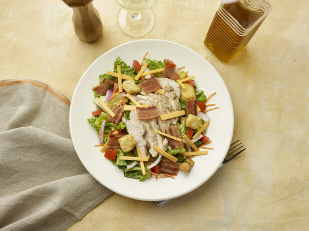 Overhead view of salad with bacon, chicken, red onion, croutons, red pepper and shredded cheddar with a glass of white wine on a beige background