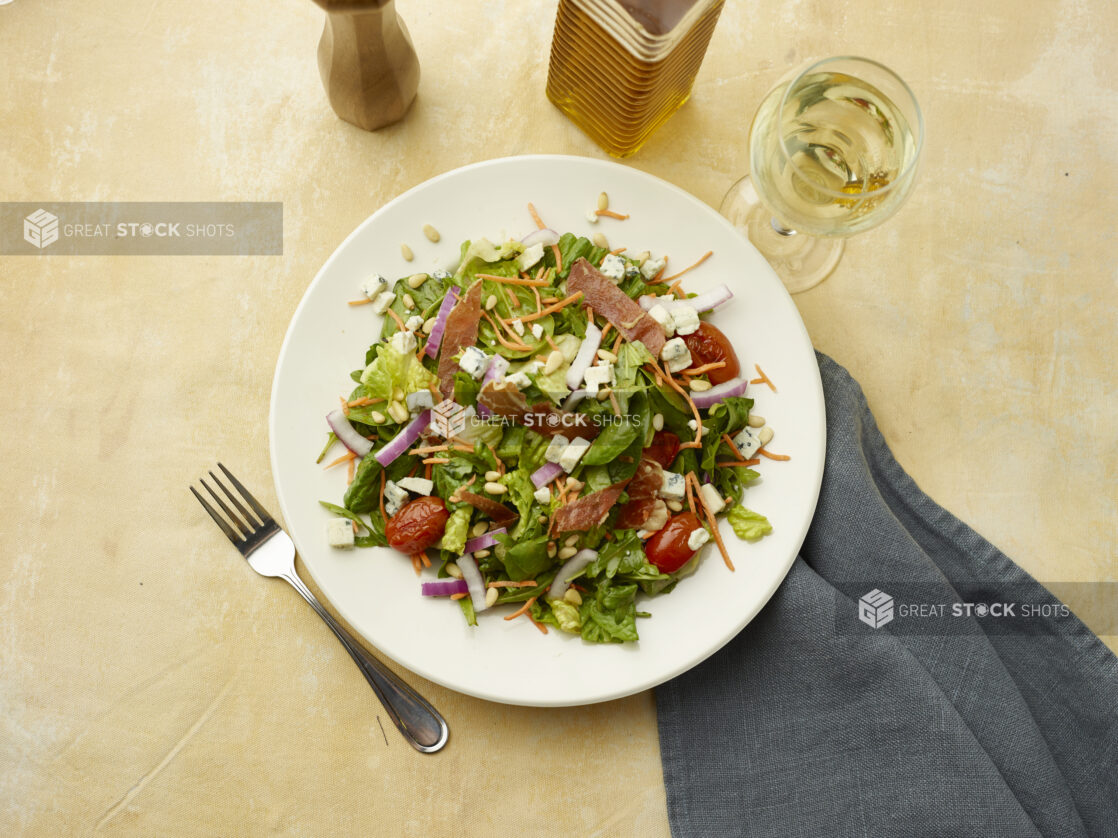 Overhead view of salad with bacon, blistered cherry tomatoes, red onion, blue cheese and pine nuts with a glass of white wine on a beige background