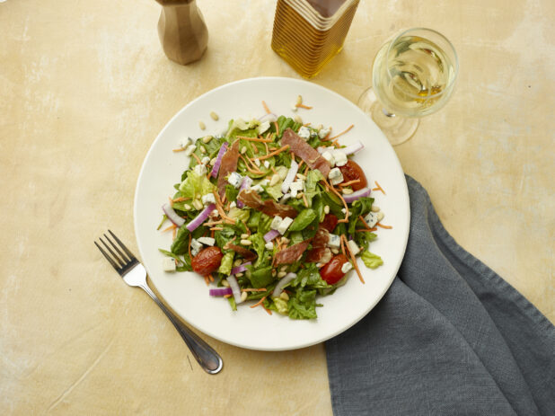 Overhead view of salad with bacon, blistered cherry tomatoes, red onion, blue cheese and pine nuts with a glass of white wine on a beige background