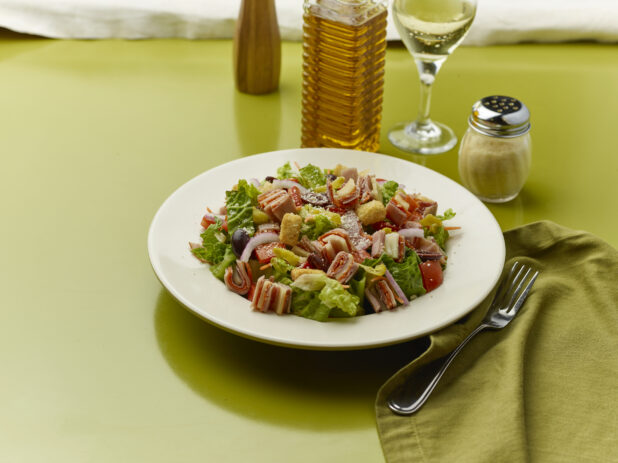 Chopped salad with sliced ham, salami and provolone squares on top of red onion, black olives and croutons with a glass of white wine on a green background