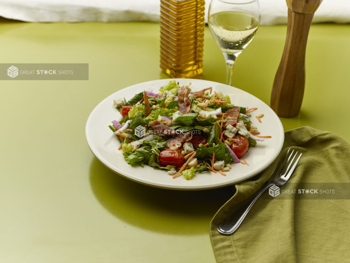 Chopped salad with bacon, blistered cherry tomatoes, red onion, blue cheese and pine nuts with a glass of white wine on a green background