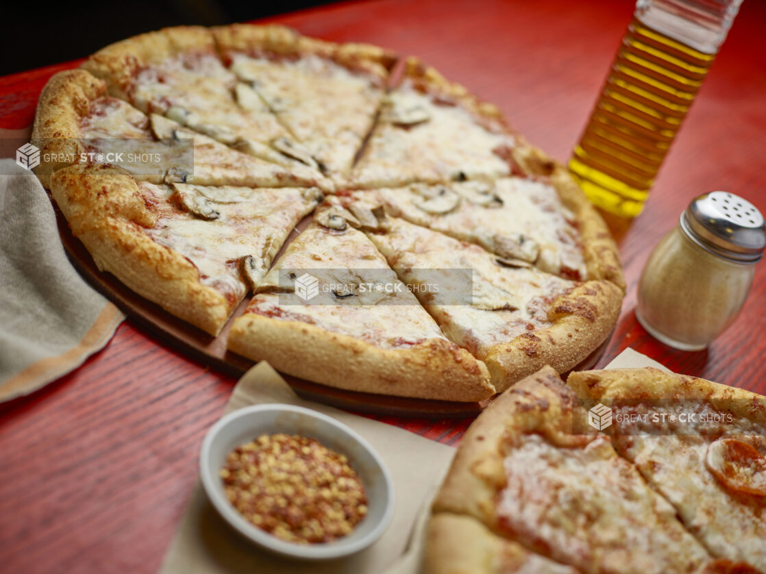 Two sliced medium pizzas with one topping with toppings in the background on a red wooden table