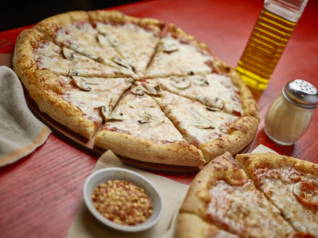 Two sliced medium pizzas with one topping with toppings in the background on a red wooden table