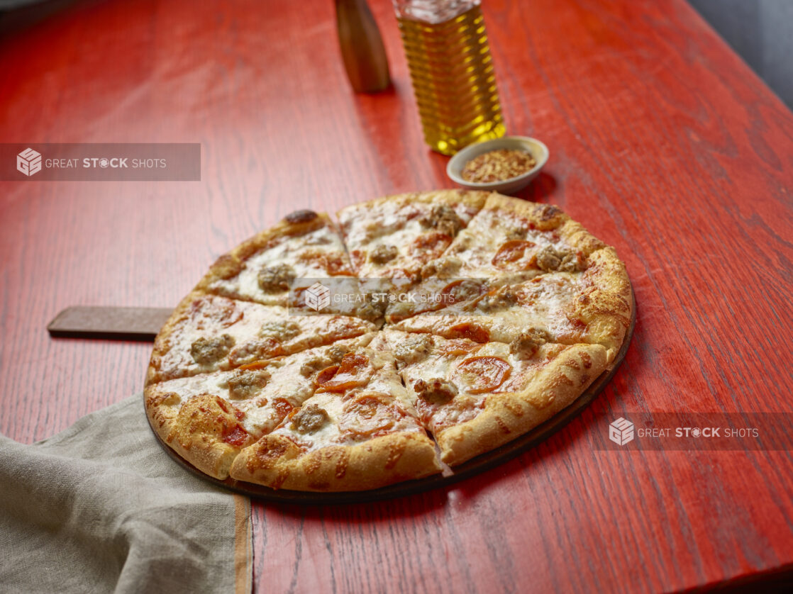 Sliced medium pizza with pepperoni and sausage with toppings in the background on a red wooden table